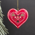 A red heart-shaped metal hanging ornament with hand-painted white, green, and gold floral and geometric patterns, hanging from a gold string against a dark background.