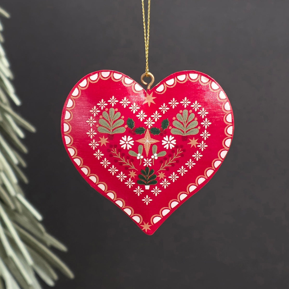 A red heart-shaped metal hanging ornament with hand-painted white, green, and gold floral and geometric patterns, hanging from a gold string against a dark background.