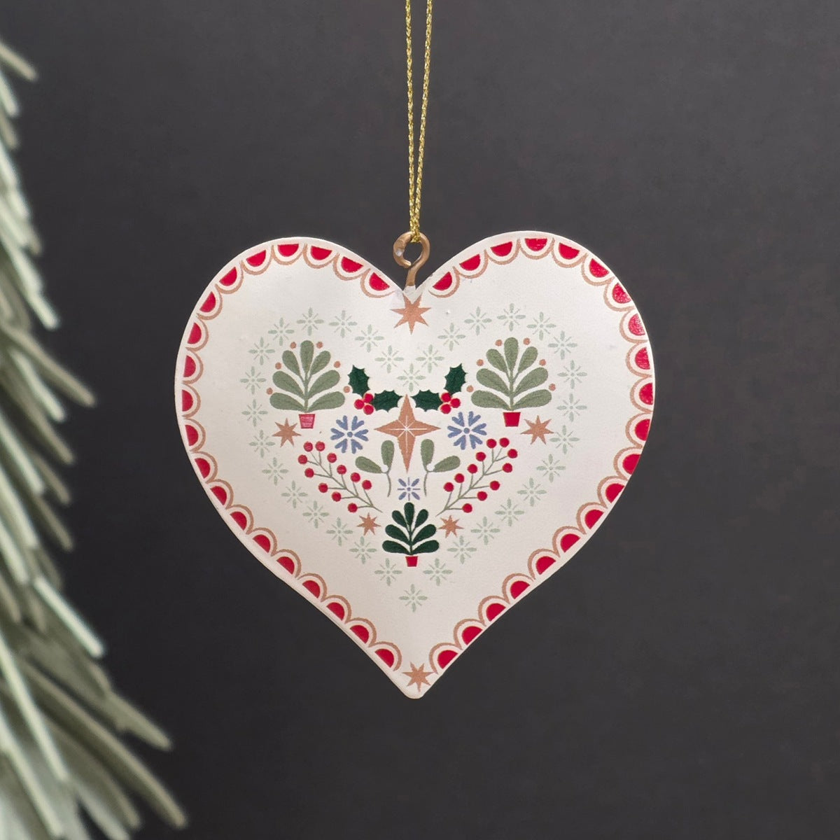 A white heart-shaped metal hanging ornament with hand-painted white, red, green, and gold floral and geometric patterns, hanging from a gold string against a dark background.