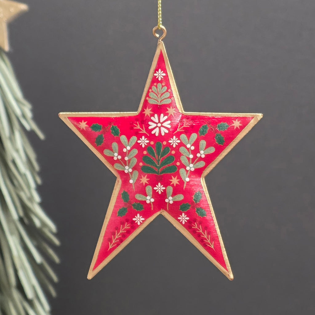 A red five-pointed metal star Christmas hanging ornament with a gold-toned edge and hand-painted green, white, and red holly and floral motifs, hanging against a dark grey background with a blurred pine branch on the left.
