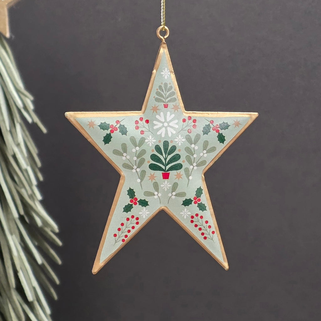 A five-pointed metal star Christmas hanging ornament with a light sage green background, decorated with a botanical pattern of holly leaves, berries, pine trees, and snowflakes, outlined in gold and hanging from a gold string.