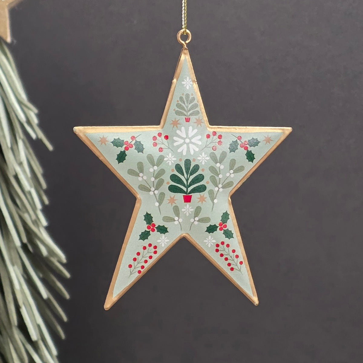 A five-pointed metal star Christmas hanging ornament with a light sage green background, decorated with a botanical pattern of holly leaves, berries, pine trees, and snowflakes, outlined in gold and hanging from a gold string.