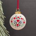 A round ceramic white Christmas ornament with a hand-painted folk art floral and heart pattern in red, green, and blue, hanging from a red ribbon against a dark grey background.
