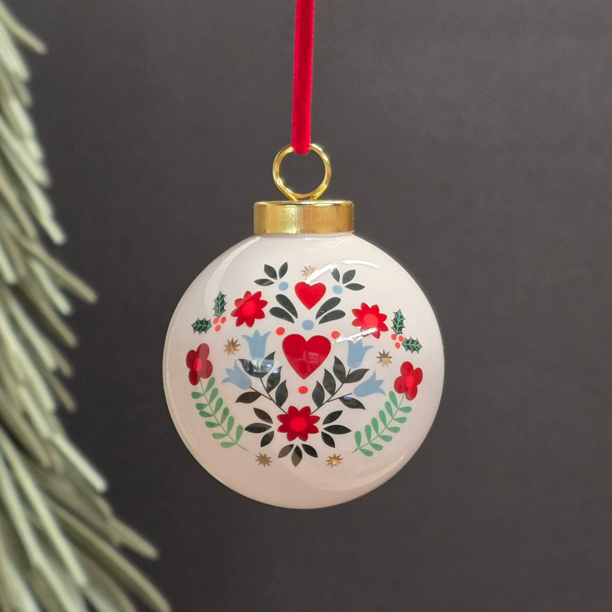 A round ceramic white Christmas ornament with a hand-painted folk art floral and heart pattern in red, green, and blue, hanging from a red ribbon against a dark grey background.