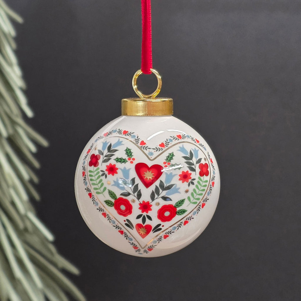 A round white ceramic Christmas ornament with a hand-painted floral heart design in red, green, and blue, hanging from a red ribbon with a gold cap on a dark background.