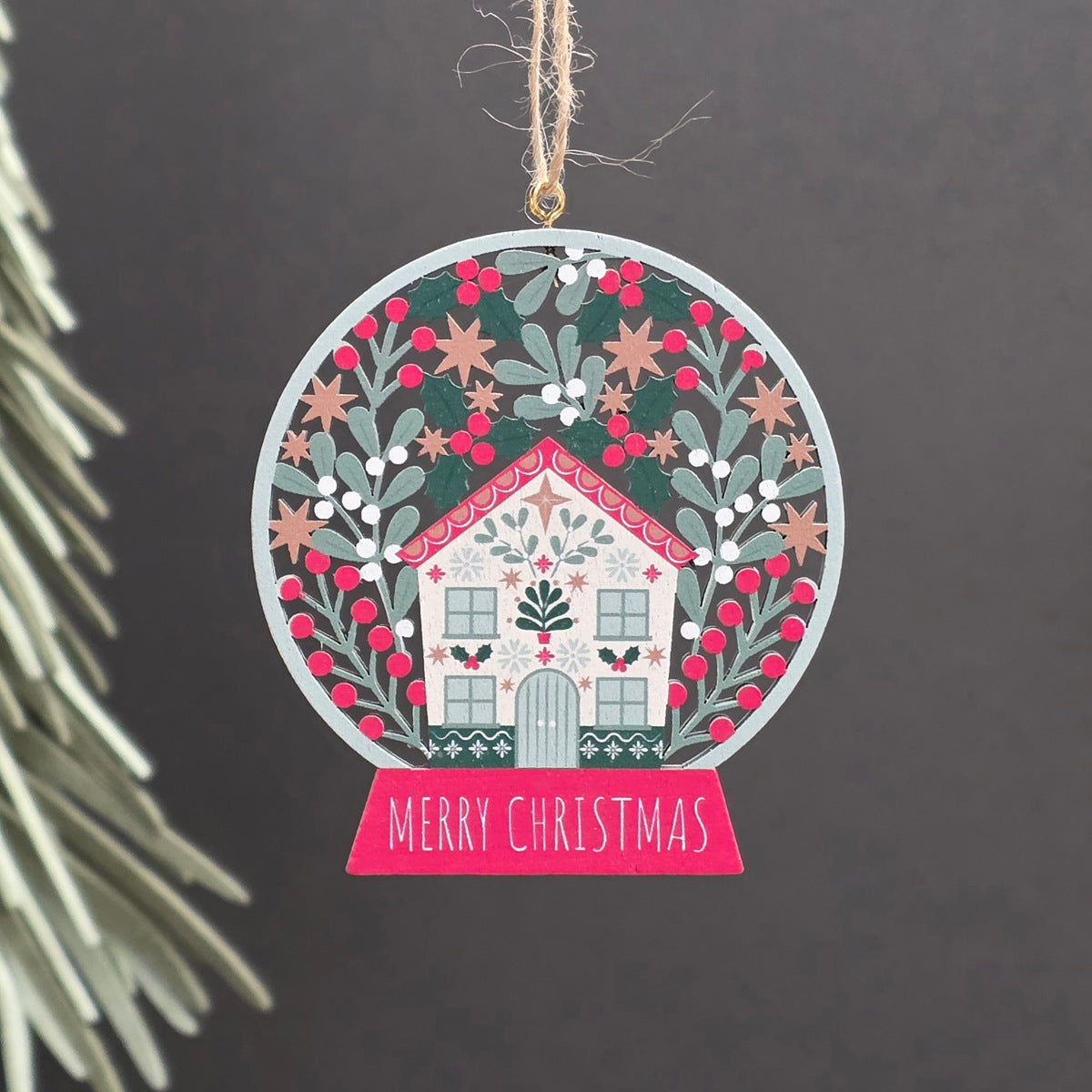 A round wooden hanging Christmas ornament featuring a small white house surrounded by green holly leaves, red berries, and gold stars, with a pink banner reading “MERRY CHRISTMAS” at the bottom. The ornament hangs from a jute string against a dark grey background with a blurred pine branch on the left.