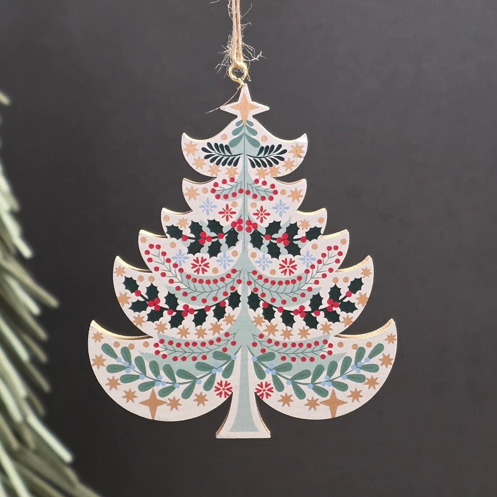 A white tree shaped wooden hanging Christmas ornament featuring green holly leaves, red berries, and gold stars.  The ornament hangs from a jute string against a dark grey background with a blurred pine branch on the left.
