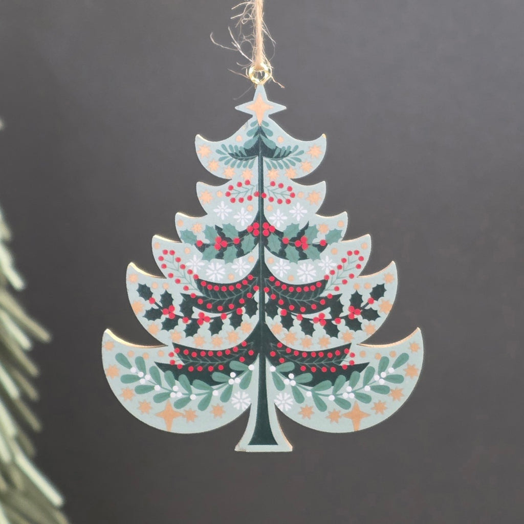 A light green tree shaped wooden hanging Christmas ornament featuring green holly leaves, red berries, and gold stars.  The ornament hangs from a jute string against a dark grey background with a blurred pine branch on the left.