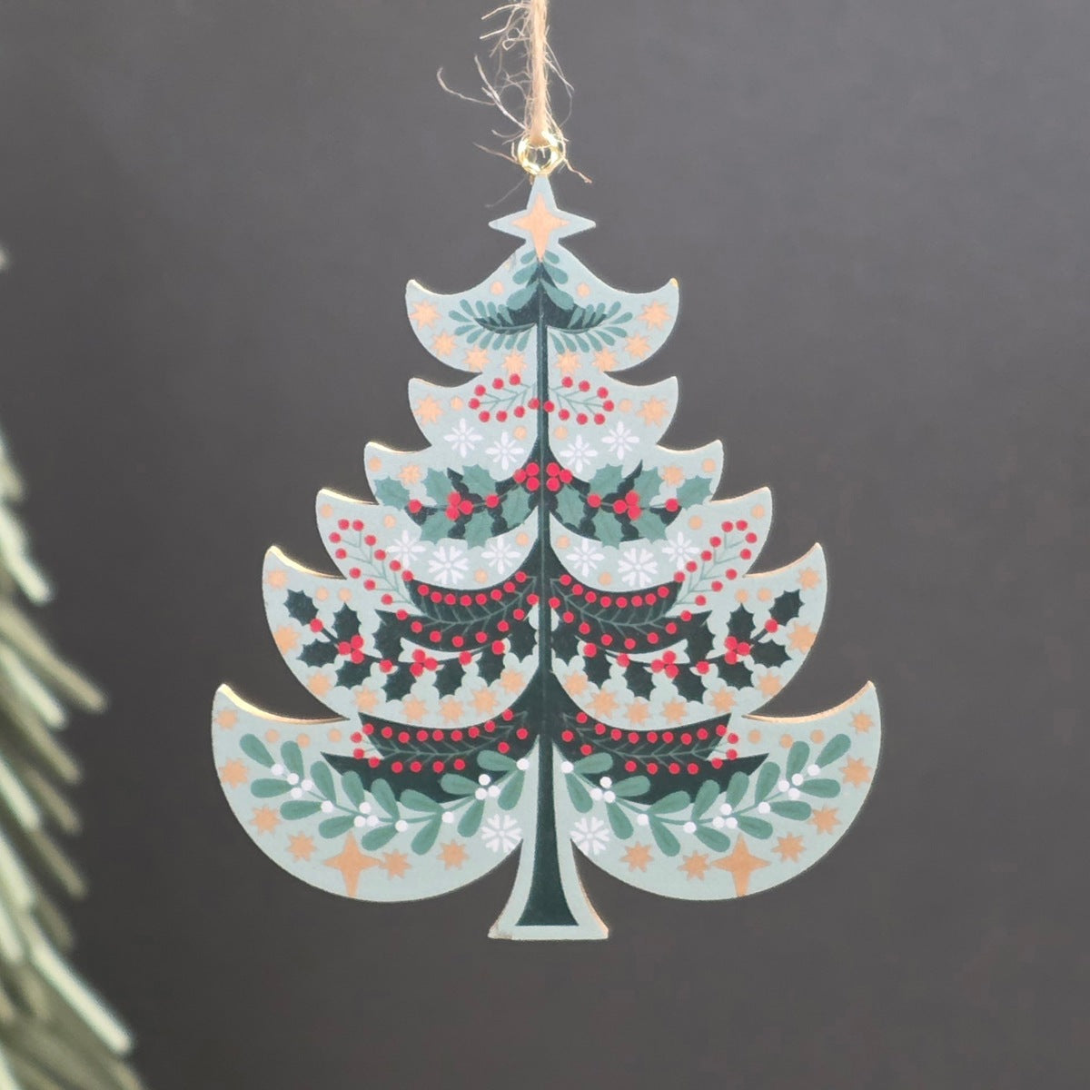 A light green tree shaped wooden hanging Christmas ornament featuring green holly leaves, red berries, and gold stars.  The ornament hangs from a jute string against a dark grey background with a blurred pine branch on the left.