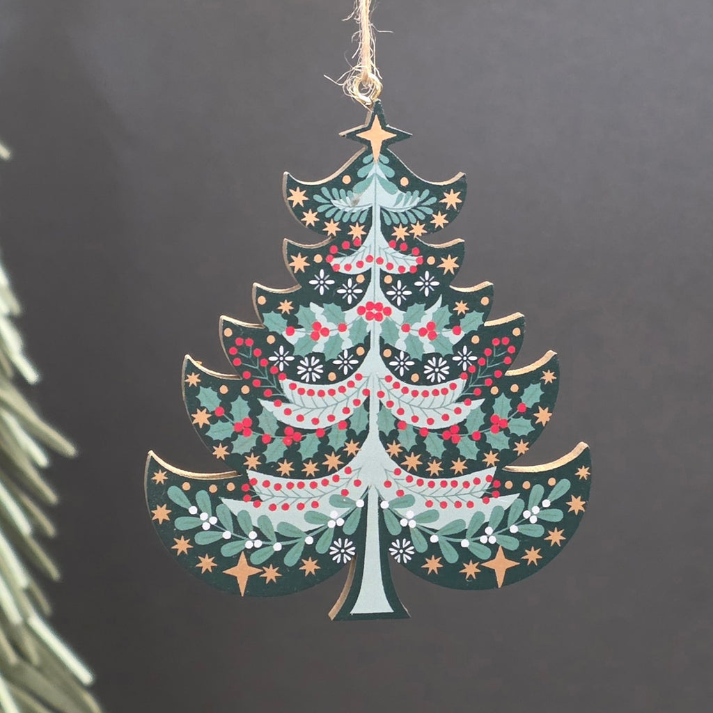 A green tree shaped wooden hanging Christmas ornament featuring green holly leaves, red berries, and gold stars.  The ornament hangs from a jute string against a dark grey background with a blurred pine branch on the left.