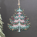 A green tree shaped wooden hanging Christmas ornament featuring green holly leaves, red berries, and gold stars.  The ornament hangs from a jute string against a dark grey background with a blurred pine branch on the left.