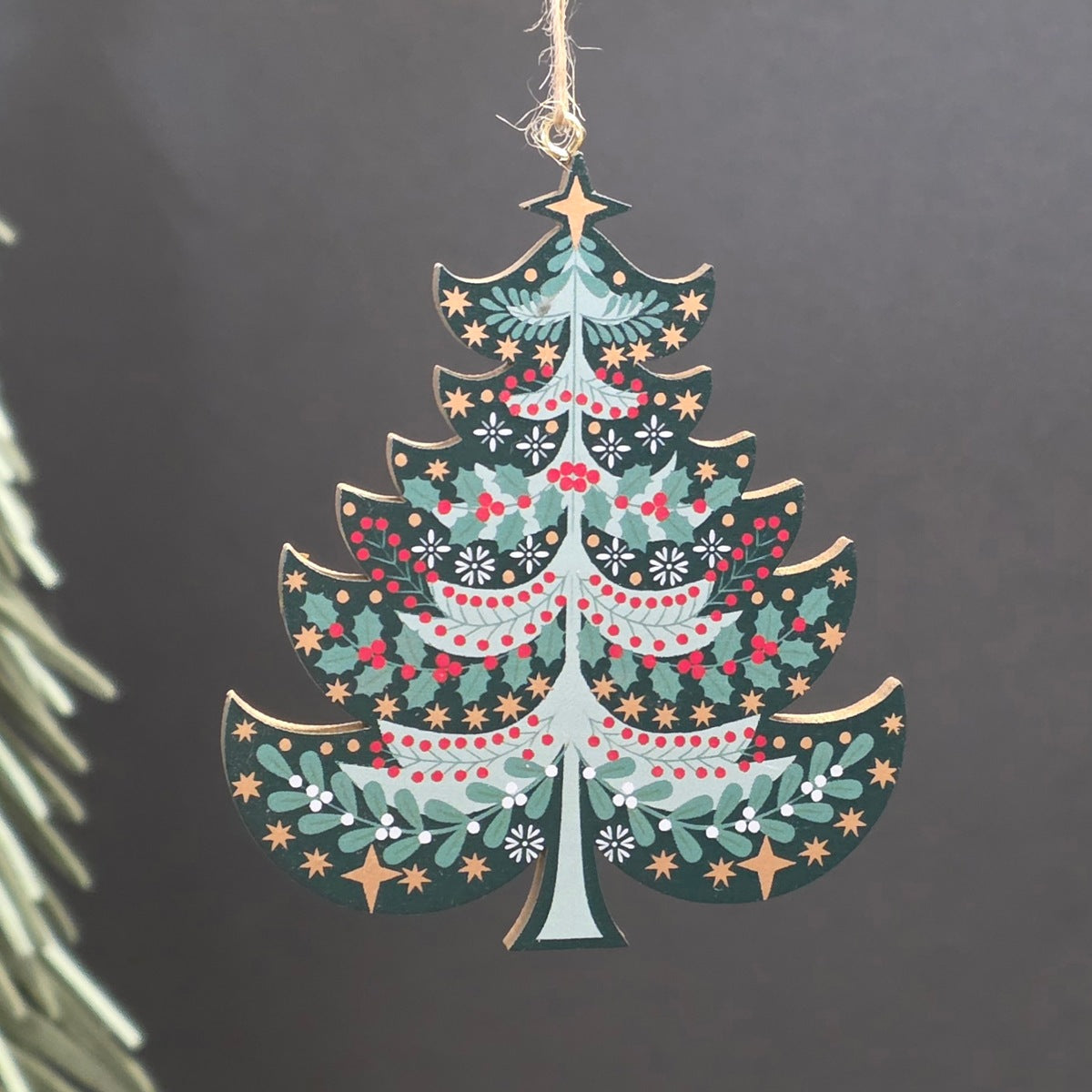A green tree shaped wooden hanging Christmas ornament featuring green holly leaves, red berries, and gold stars.  The ornament hangs from a jute string against a dark grey background with a blurred pine branch on the left.