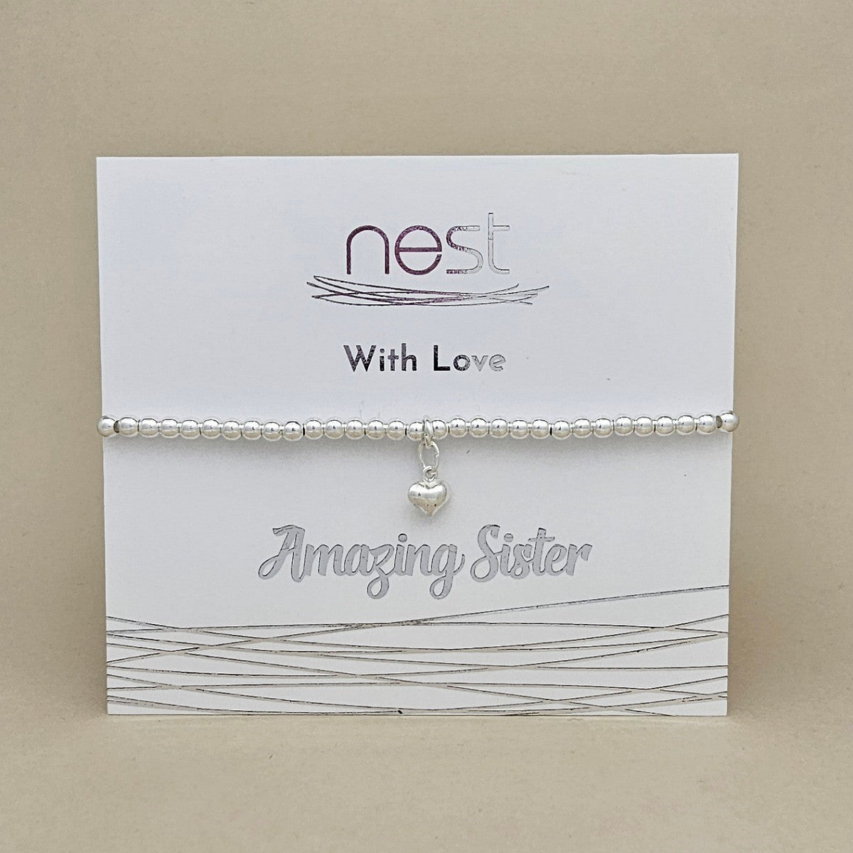 A silver beaded bracelet with a small heart charm displayed on a white card labeled 'nest With Love Amazing Sister' against a beige background.