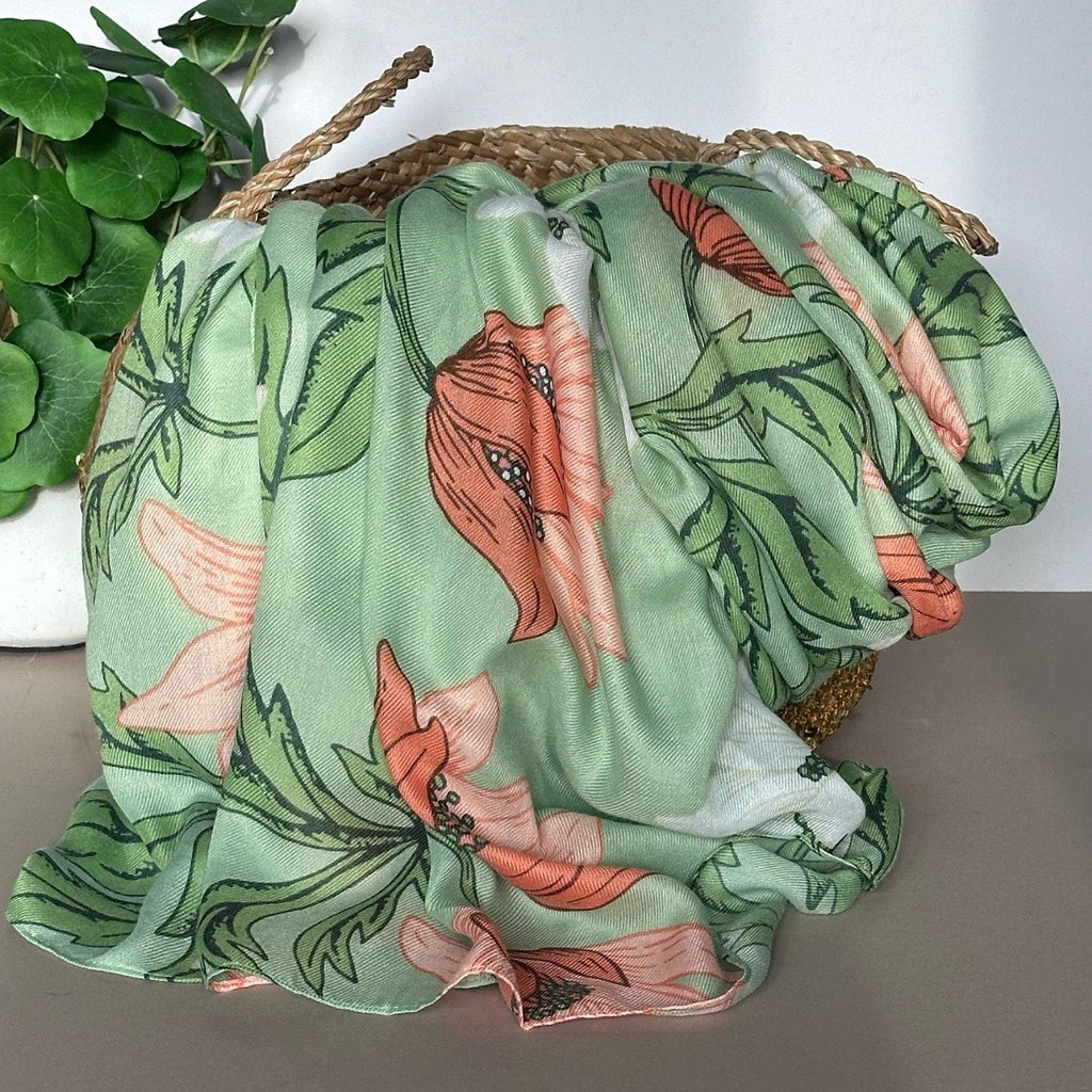 A mint green anemone floral scarf with coral, peach and green botanical patterns draped over a woven straw basket, with a green potted plant in the background.