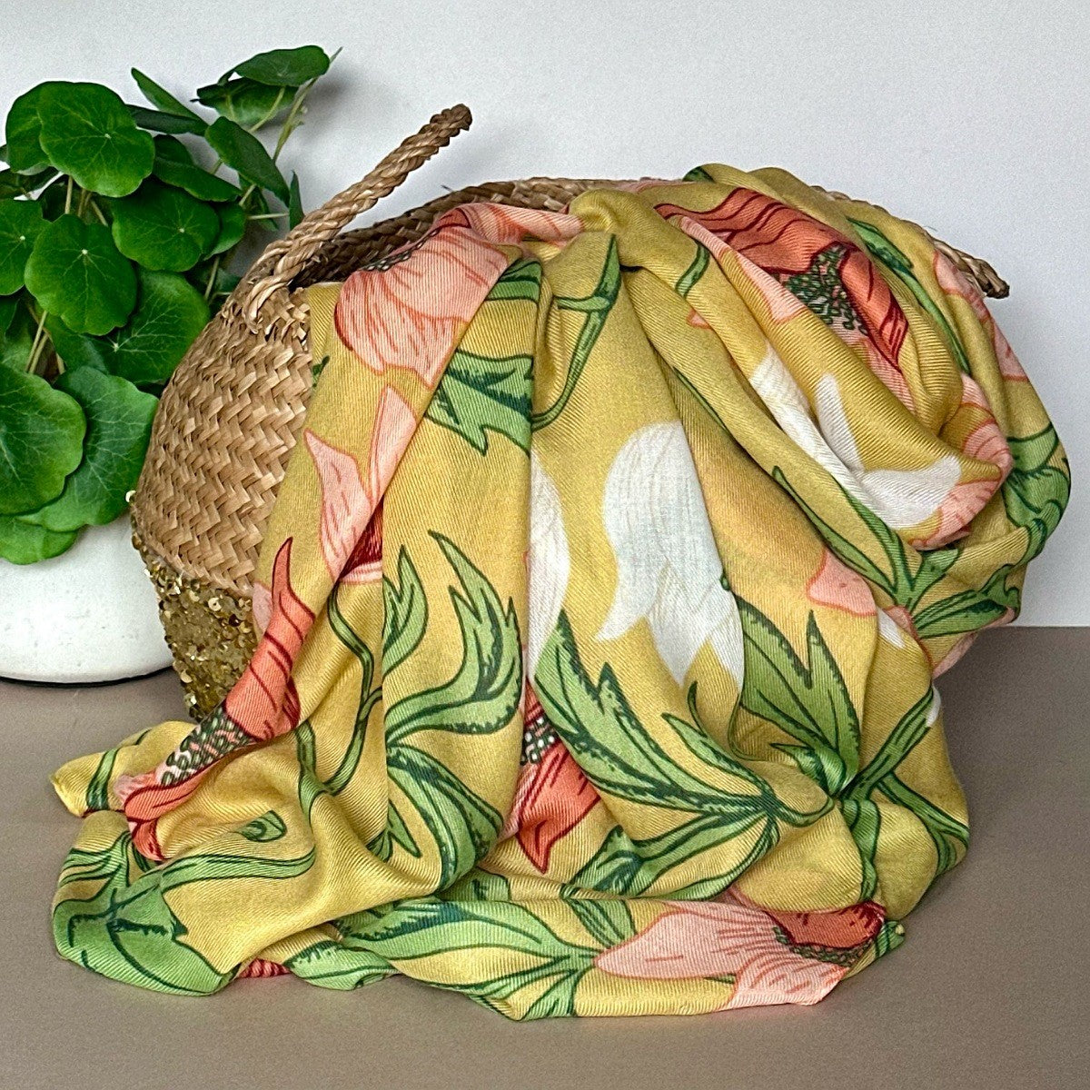 A yellow anemone floral scarf with pink, white, and green botanical patterns draped over a woven straw basket, with a green potted plant in the background.