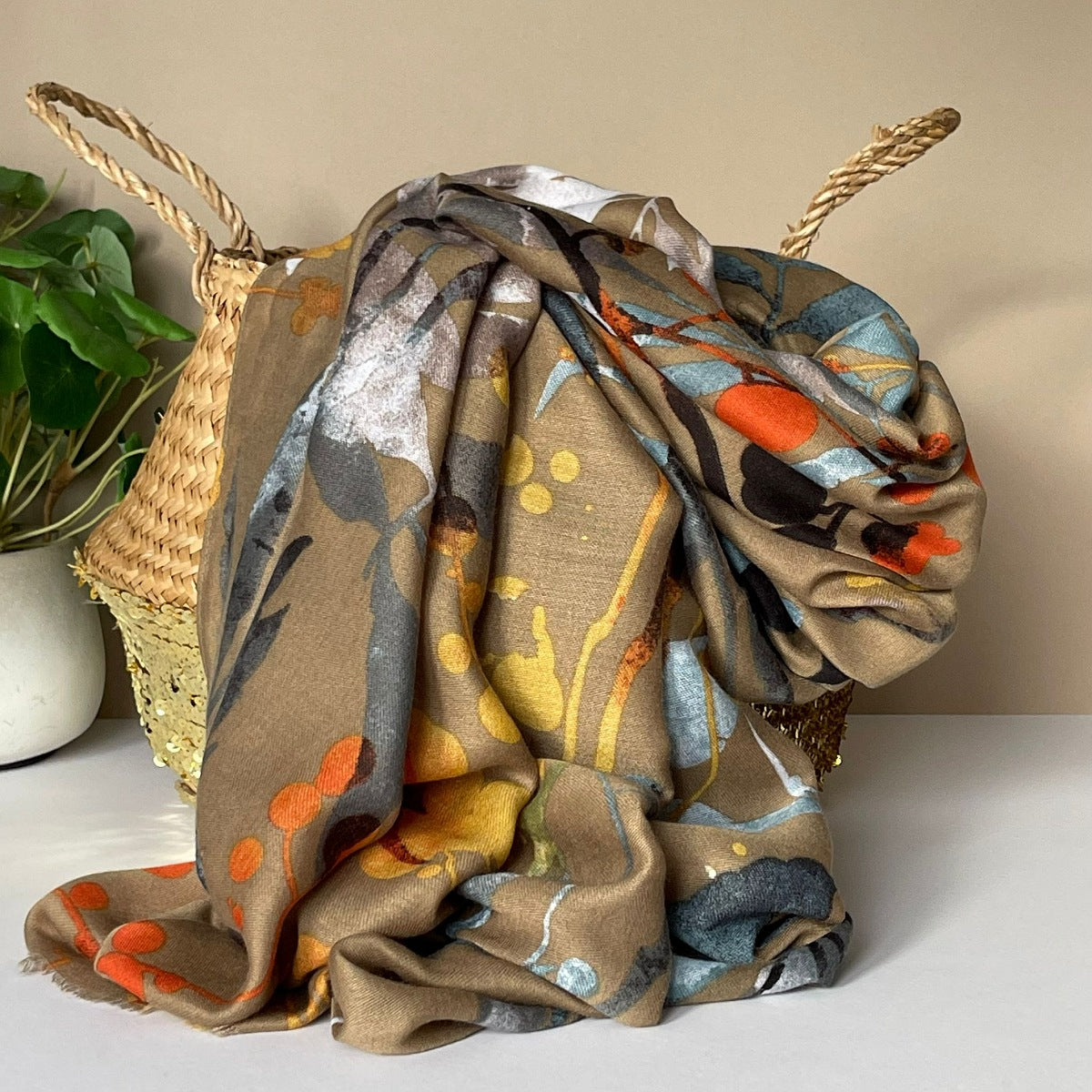 A beige scarf with autumn leaves print in mustard, orange, yellow, brown and greys, draped over a woven straw basket next to a potted green plant.
