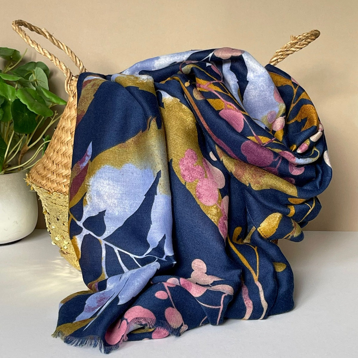 A navy scarf with autumn leaves print in mustard, pink shades, purple and violet, draped over a woven straw basket next to a potted green plant.
