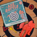 Axe Battle board game set shown with the teal game box and several bright orange axe pieces on a wooden tabletop with a black target board.