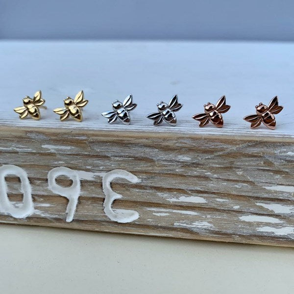 Six small bee-shaped stud earrings in gold, silver, and rose gold tones are arranged on a weathered wooden block with white painted numbers.