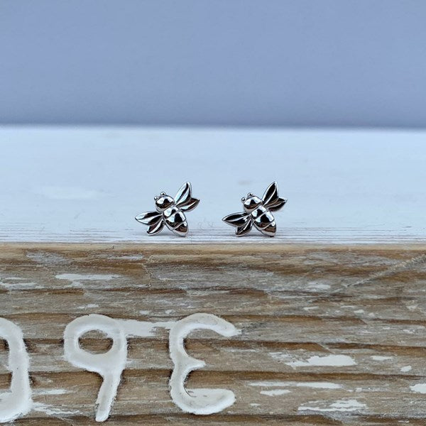 A pair of silver bee stud earrings resting on a rustic wooden surface with a light blue-gray background.