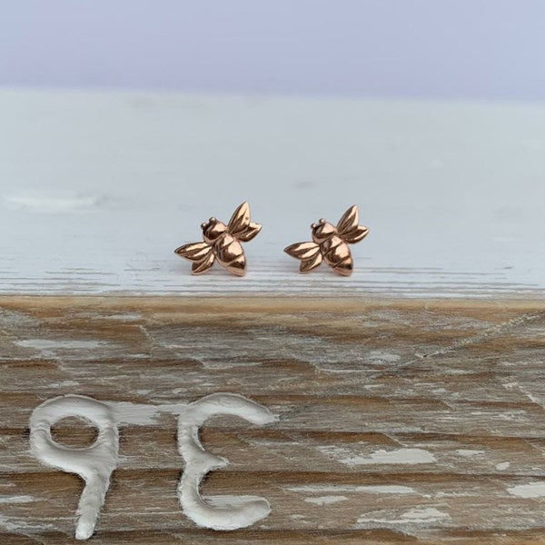 A pair of small rose gold bee stud earrings resting on a rustic wooden surface with a blurred light background.