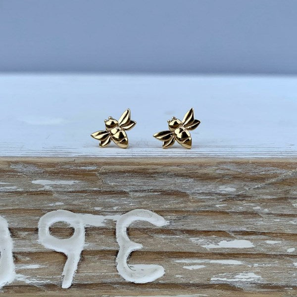 A pair of small gold bee stud earrings resting on a rustic wooden surface with a soft blue-gray background.