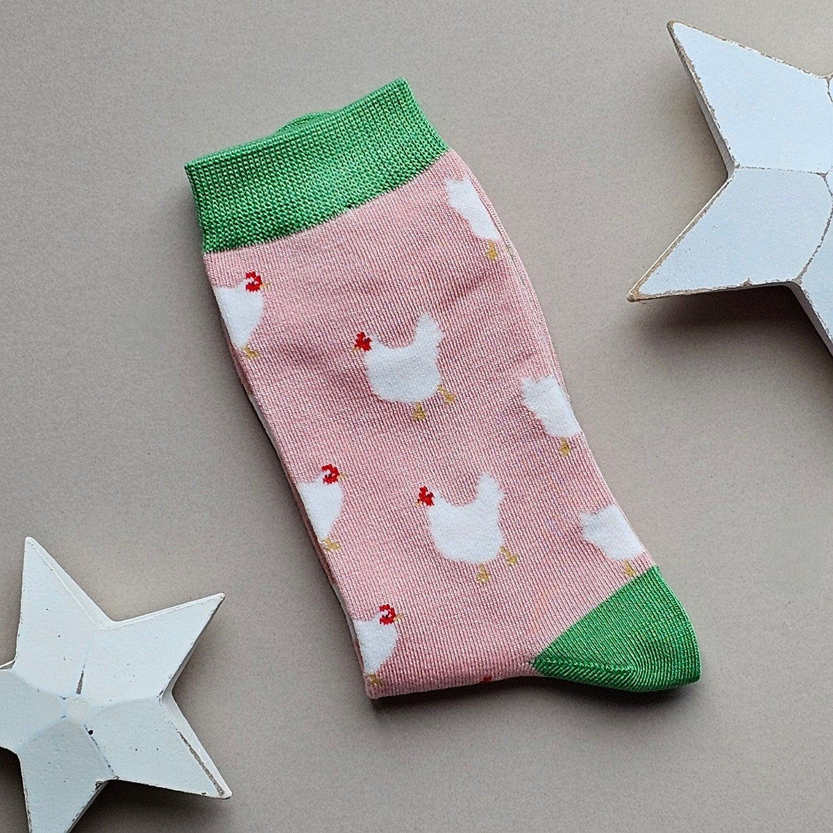 A single light pink sock with numerous white chicken patterns and green cuffs, heel and toe, laid flat on a neutral background with white star props.