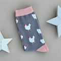 A single grey sock with numerous white chicken patterns and light pink cuffs, heel and toe, laid flat on a neutral background with white star props.
