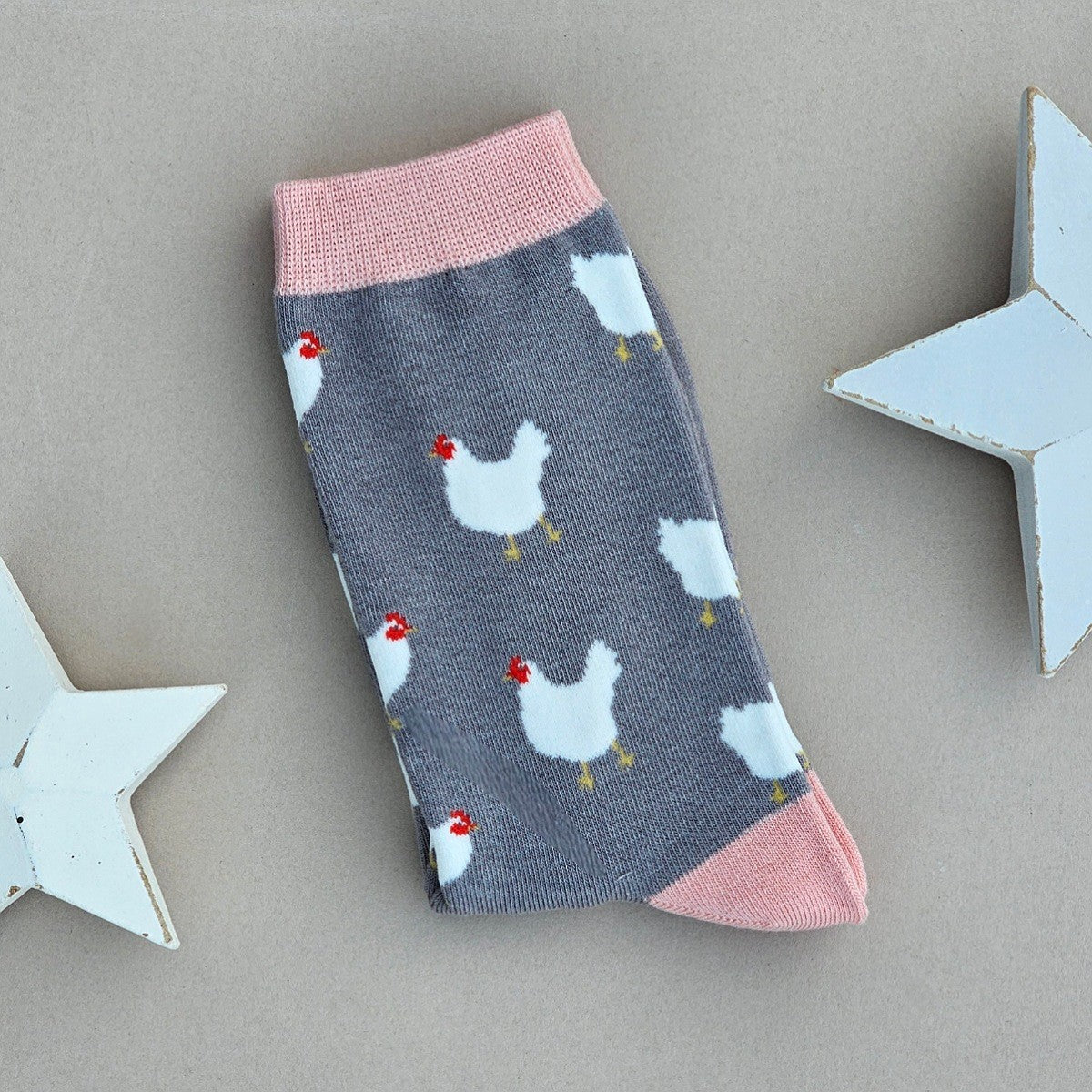 A single grey sock with numerous white chicken patterns and light pink cuffs, heel and toe, laid flat on a neutral background with white star props.