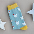 A single light blue sock with numerous white chicken patterns and yellow cuffs, heel and toe, laid flat on a neutral background with white star props.