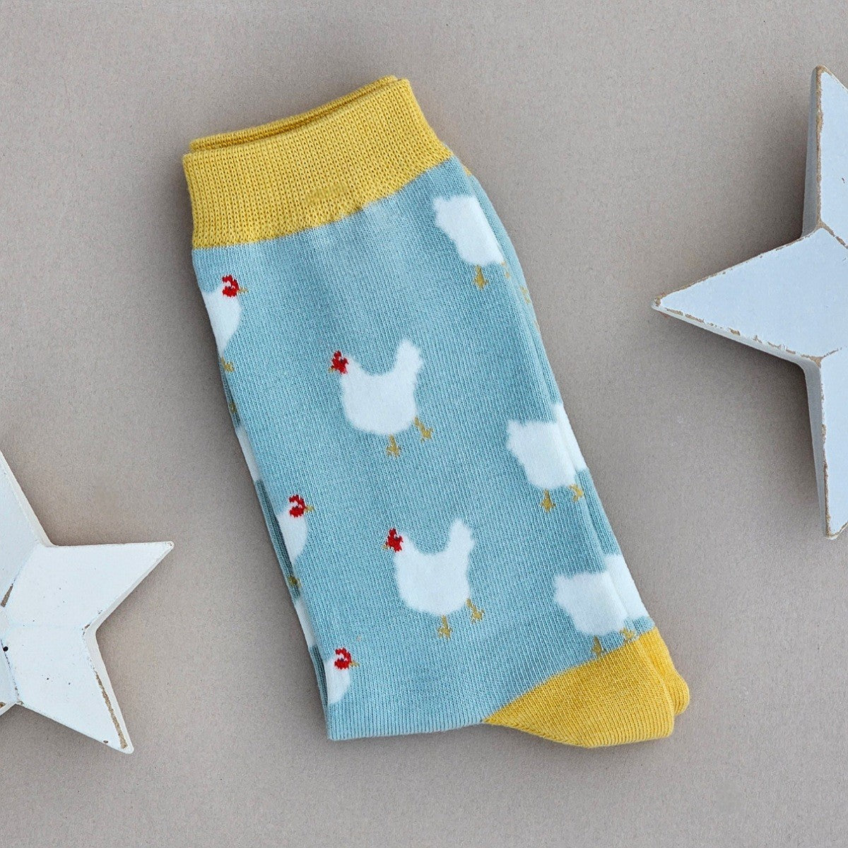 A single light blue sock with numerous white chicken patterns and yellow cuffs, heel and toe, laid flat on a neutral background with white star props.
