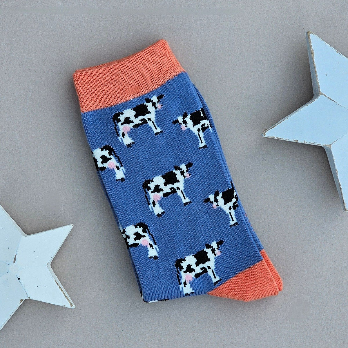 A single blue sock with a black-and-white cow print, coral cuff, heel and toe.  Laid flat on a neutral background with decorative white stars.