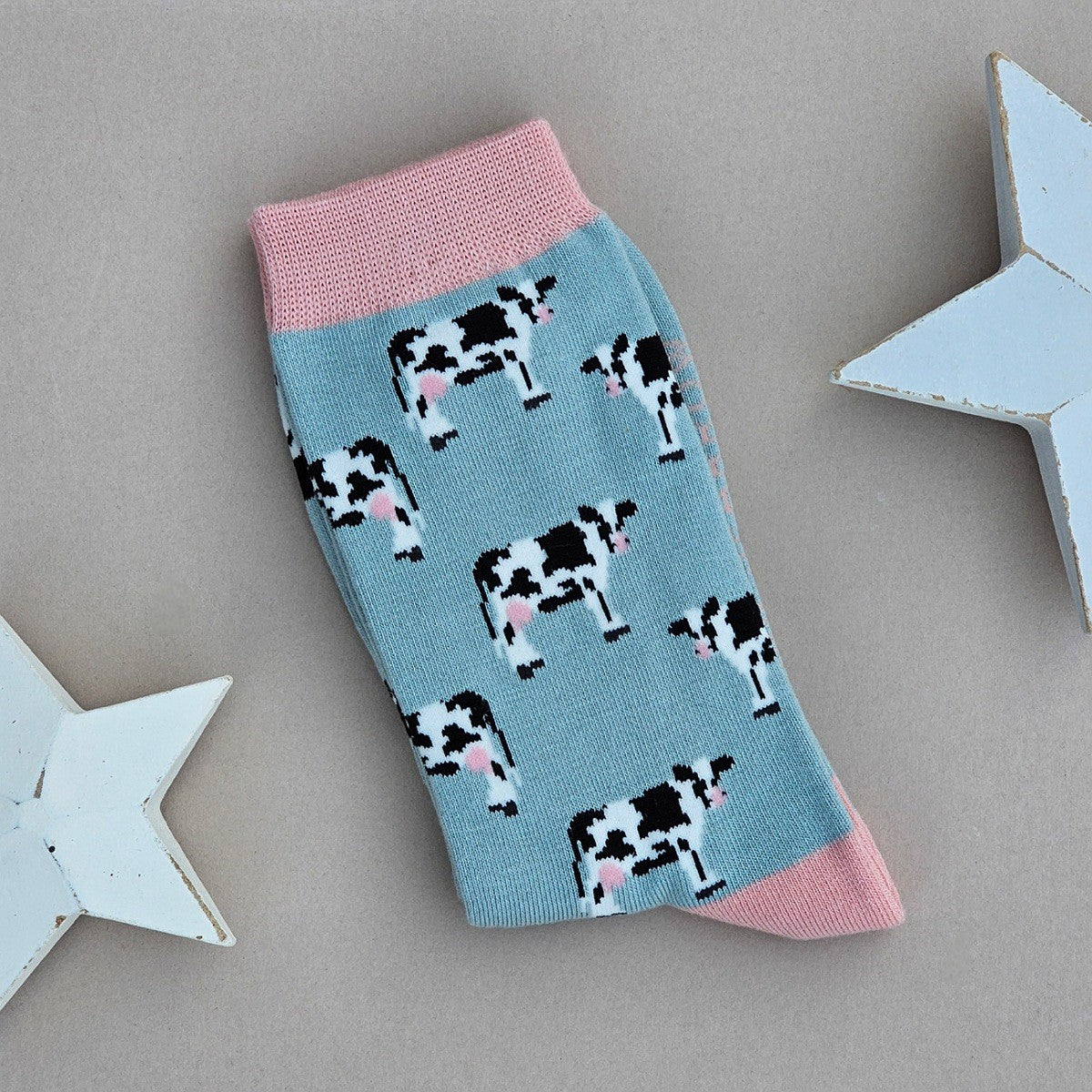 A single duck egg blue sock with a black-and-white cow print, light pink cuff, heel and toe.  Laid flat on a neutral background with decorative white stars.