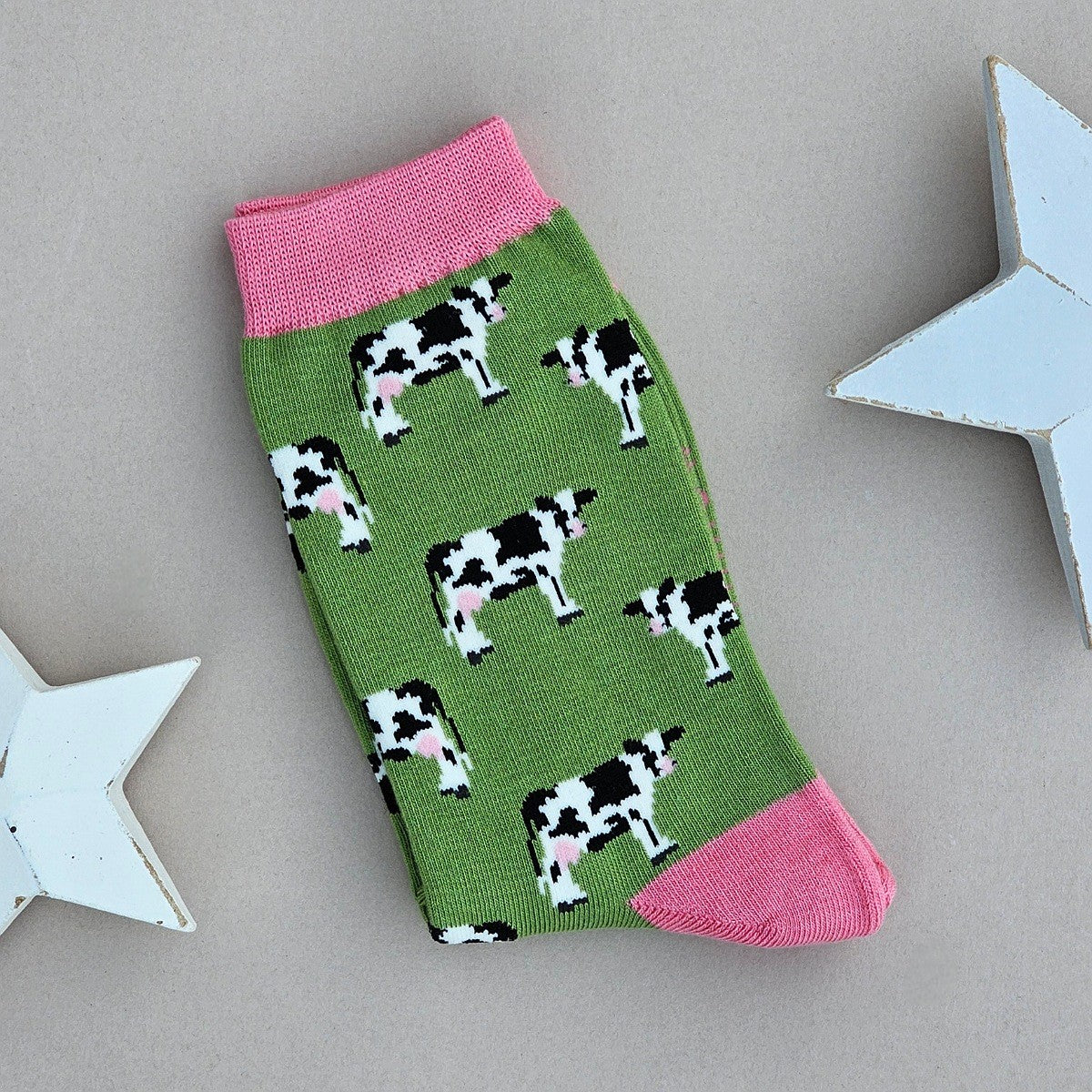 A single green sock with a black-and-white cow print, pink cuff, heel and toe.  Laid flat on a neutral background with decorative white stars.