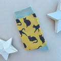 A single yellow sock with numerous black cat patterns and light blue ribbed cuff, heel and toes.  Displayed on a light grey surface with white star-shaped props.