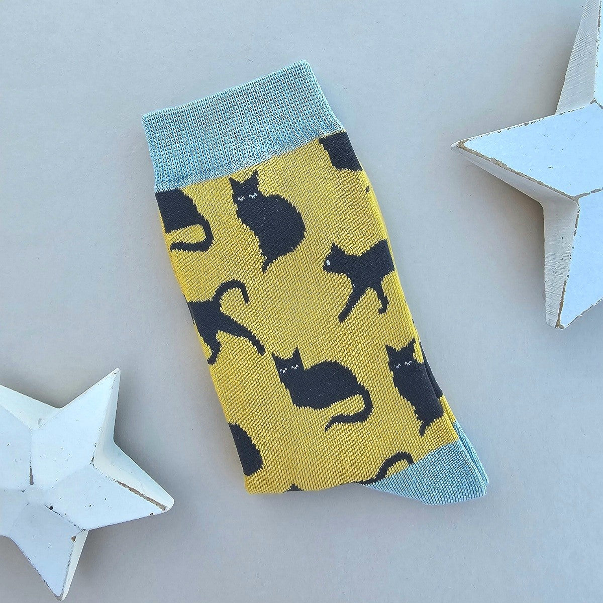A single yellow sock with numerous black cat patterns and light blue ribbed cuff, heel and toes.  Displayed on a light grey surface with white star-shaped props.