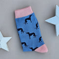 A single sock laid flat on a light beige background, decorated with black grey hound dog silhouettes on a blue base, with pink ribbed cuffs, toe and heels. Decorative white stars are placed on both sides.
