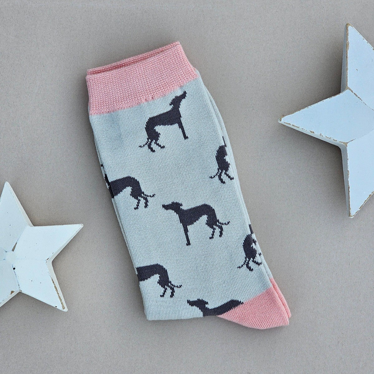 A single sock laid flat on a neutral grey background, decorated with black grey hound dog silhouettes on a silver base, with pink ribbed cuffs, toe and heels. Decorative white stars are placed on both sides.