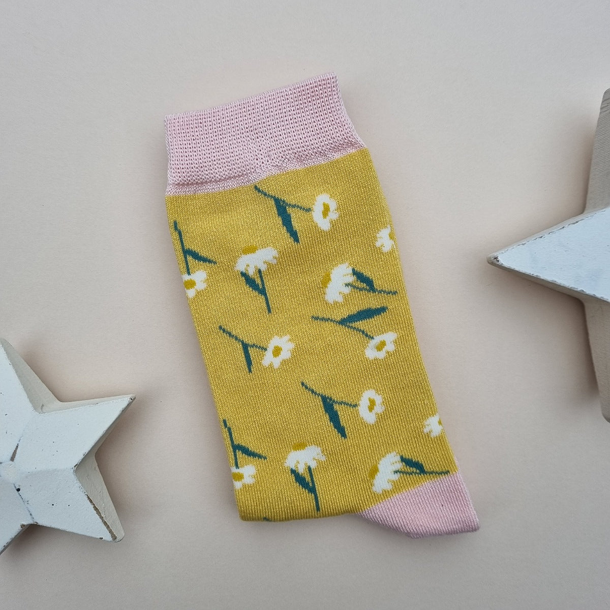 A single yellow knit sock with a white daisy and green stem pattern, featuring light pink ribbed cuffs, heel and toe, displayed on a neutral beige background with decorative white star props.