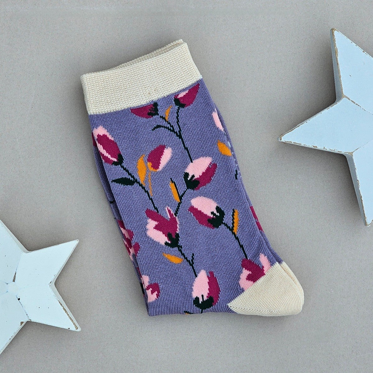 A single purple sock with a light and dark pink floral pattern, featuring beige ribbed cuffs, heel and toe, displayed on a neutral grey background with decorative white stars.
