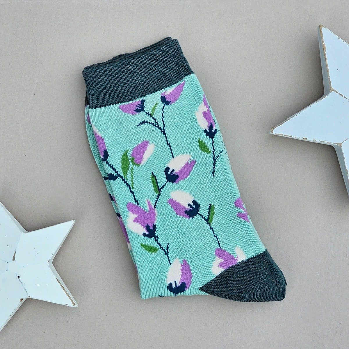 A single mint green sock with a purple and white floral pattern, featuring dark green ribbed cuffs, heel and toe, displayed on a neutral grey background with decorative white stars.