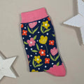 A single knit sock laid flat on a beige surface, featuring a dark navy background with a colourful floral pattern in pink, yellow, orange, and green, plus pink ribbed cuffs and heels. Decorative white star props are partially visible around the edges.