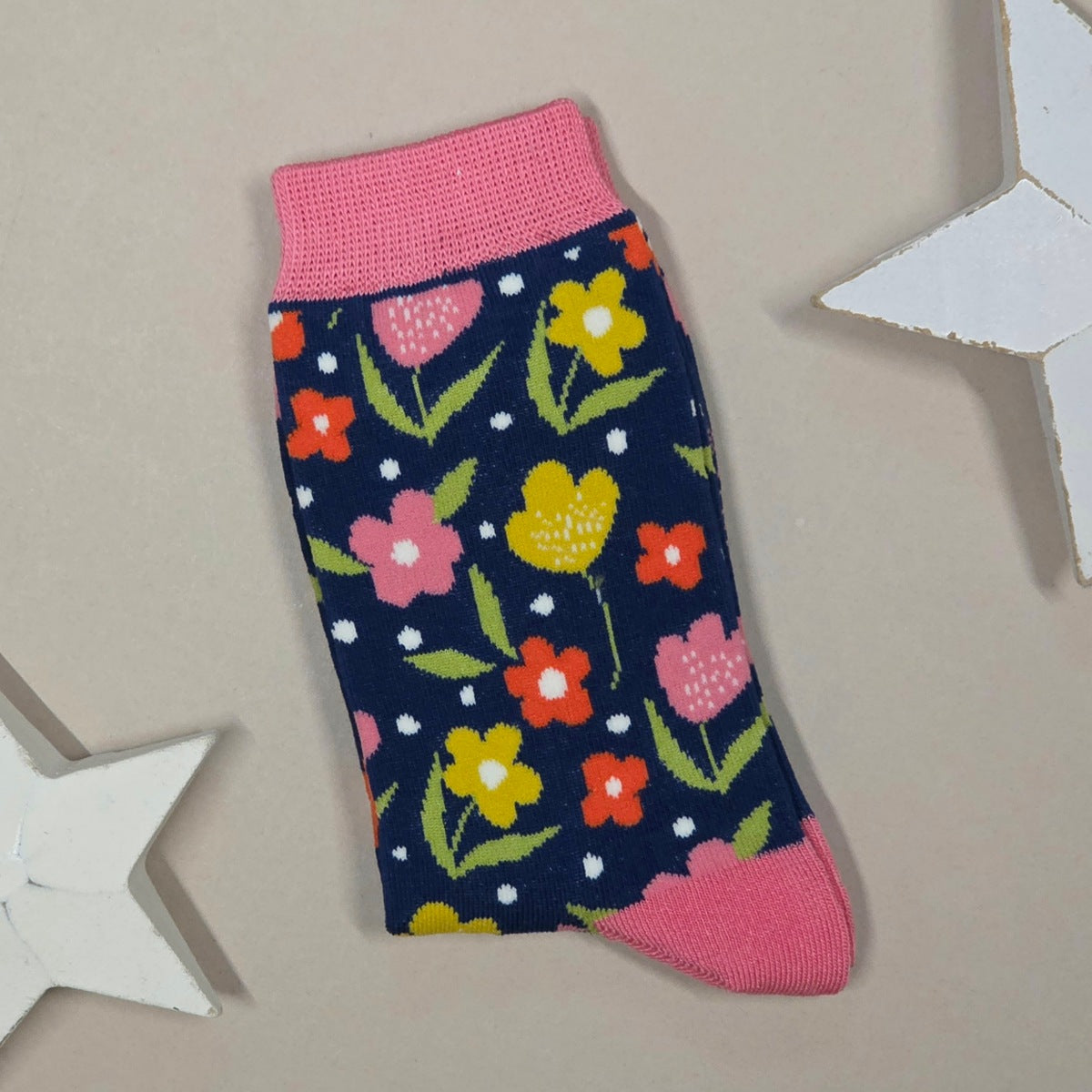 A single knit sock laid flat on a beige surface, featuring a dark navy background with a colourful floral pattern in pink, yellow, orange, and green, plus pink ribbed cuffs and heels. Decorative white star props are partially visible around the edges.