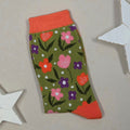 A single knit sock with a green base, orange, purple and pink floral pattern, and orange cuff and heel, displayed on a beige surface with white star-shaped props.