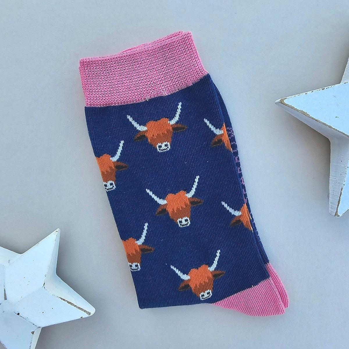 A single folded pair of socks with a navy blue base, featuring a repeating pattern of orange and white highland cow heads, finished with a pink ribbed cuff, toe/heel caps. The socks are styled on a light grey surface with white wooden star props.