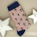 A single dusky pink sock with a repeating pattern of honey bees, featuring a navy blue cuff, heel and toe, displayed on a neutral beige background with white star-shaped props.
