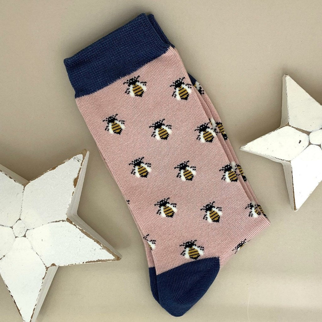 A single dusky pink sock with a repeating pattern of honey bees, featuring a navy blue cuff, heel and toe, displayed on a neutral beige background with white star-shaped props.
