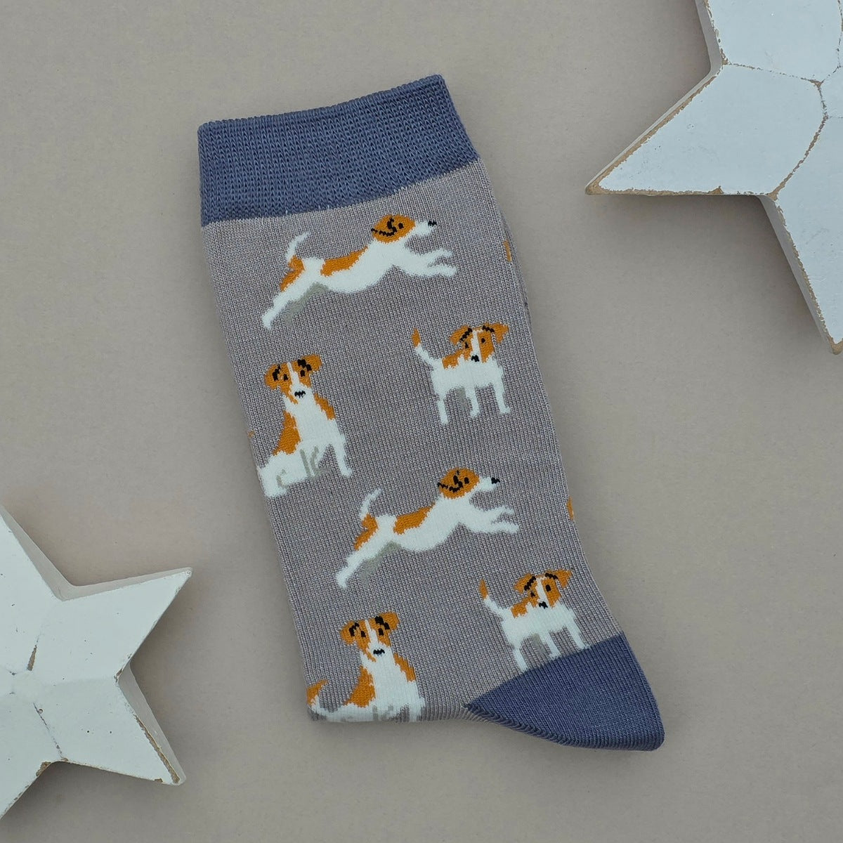 A single grey sock with a pattern of brown and white Jack Russell Terrier dogs, featuring denim blue cuffs, heel and toe, displayed on a neutral grey background with white star-shaped props.