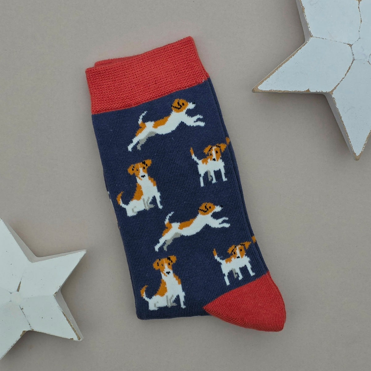 A single navy blue sock with a pattern of brown and white Jack Russell Terrier dogs, featuring red cuffs, heel and toe, displayed on a neutral grey background with white star-shaped props.