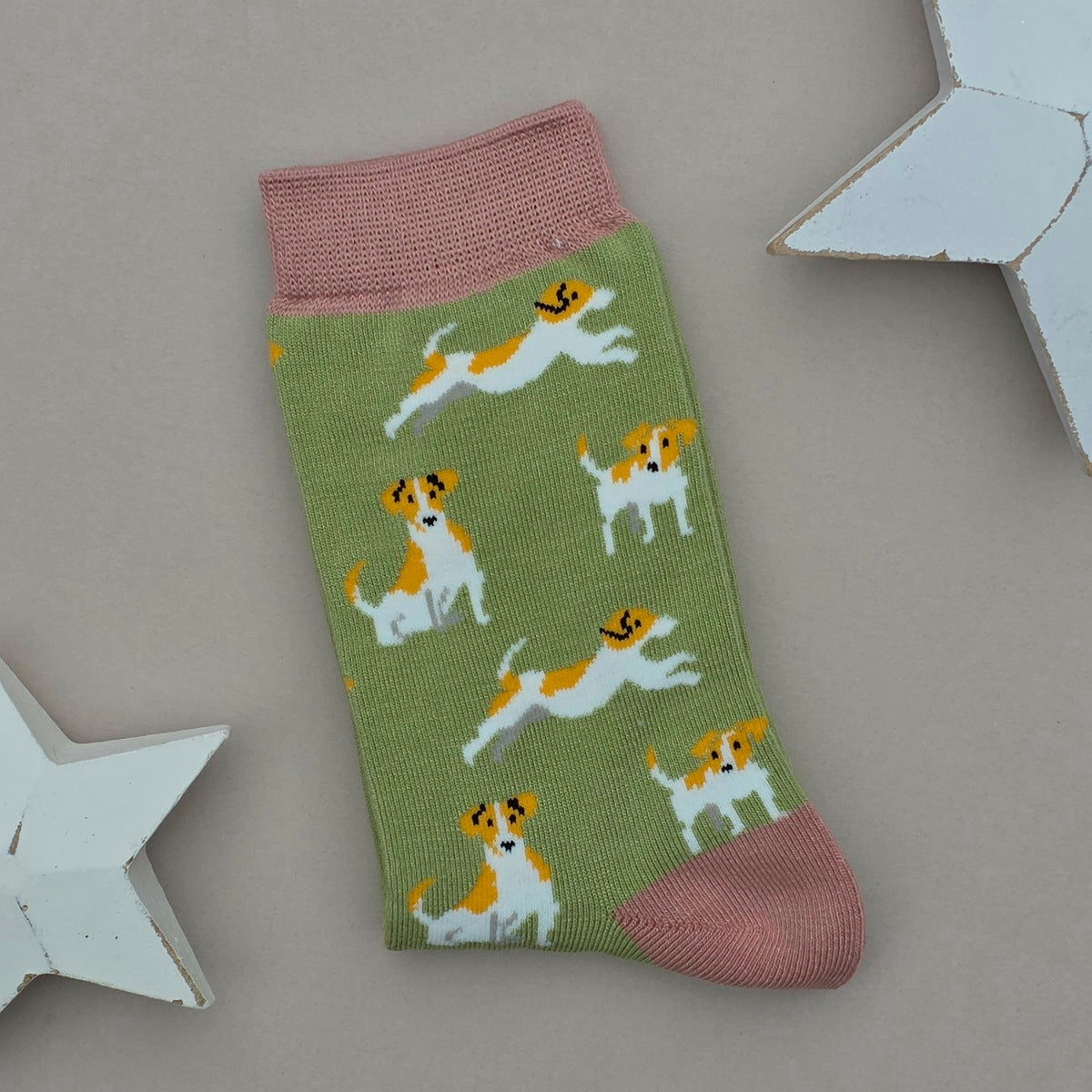 A single olive green sock with a pattern of brown and white Jack Russell Terrier dogs, featuring pink cuffs, heel and toe, displayed on a neutral grey background with white star-shaped props.